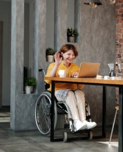 Imagen de una chica en silla de ruedas que esta sonriendo mientras saluda a la pantalla de su ordenador portátil mientras hace una videollamada.