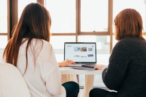 Dos chicas están sentadas delante de un ordenador portátil mirando una pagina web.