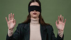 Una mujer con un jersey de cuello alto beige, una americana negra y ojos vendados con una cinta de ese mismo color, levanta ambas manos con las palmas abiertas hacia delante.