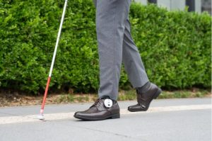 Plano corto del dispositivo Ashirase en unos zapatos acompañado de un bastón blanco.