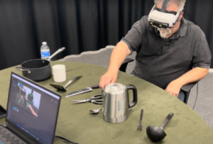 Un hombre lleva unas gafas de realidad virtual sentado en una silla frente a una mesa con diferentes utensilios de cocina: una botella de agua, una pota, unas tijeras, un cuchillo, un cazo, una cuchara y un tenedor. En el otro extremo de la mesa un portátil que enseña en su pantalla la imagen que captan las gafas del hombre, sombreando los objetos en diferentes colores acorde a su peligrosidad.