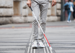 Plano corto de las piernas de un hombre cruzando un paso de peatones por el itinerario podotáctill de un paso de peatones ayudado de un bastón de sordoceguera.